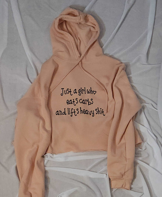 A close-up of a pink crop hoodie with the phrase "Just a Girl Who" printed on the front. The hoodie is made of super soft airlume spun cotton and features a trendy crop design.