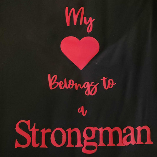 Black tee with a red cute print that says "My Heart Belongs to a Strongman." Available in ladies cut sizes small through 2X or unisex sizes small through 5X. Made from high-quality cotton and perfect for all-day wear. Option to add the print to the front